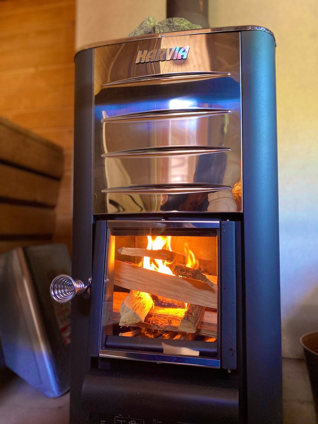 Front view of a wood-fired sauna heater, showcasing its design with a chimney and a door for loading wood, set against a wooden backdrop.