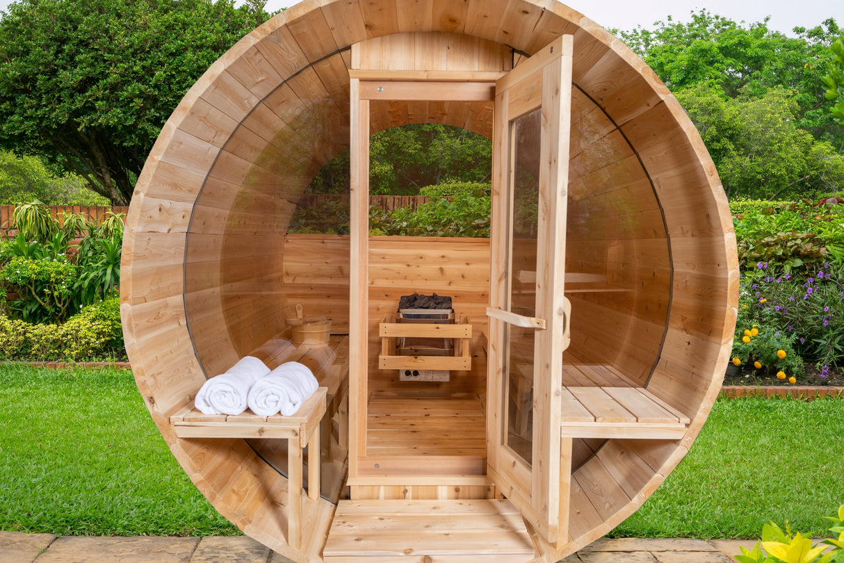 Front view of the Serenity MP Barrel Sauna with an open glass door, showcasing a sauna heater inside, along with a porch and windows.