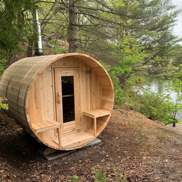 Front view of a Serenity Barrel Sauna with a porch and glass door, located in a forested area.