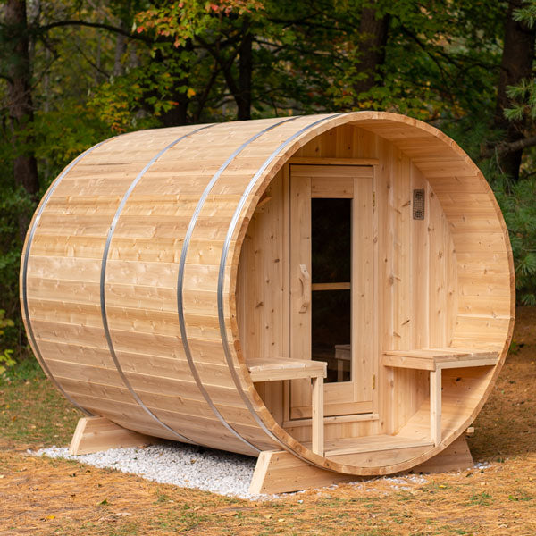 Front and left side view of a Serenity Barrel Sauna, featuring a porch and glass door, situated in a forested area.