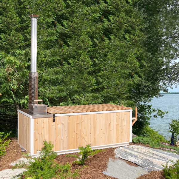 Back angle view of the Starlight Hot Tub in cedar, featuring the visible flue pipe, situated near a tree and a lake.