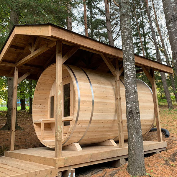 Front and side view of a tranquility barrel sauna with a porch, glass windows, and doors, all covered by a roof.