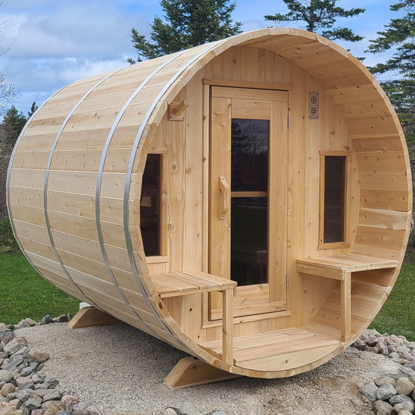 Front and left side view of a tranquility barrel sauna featuring a porch, glass windows, and doors, situated on a sandy area.