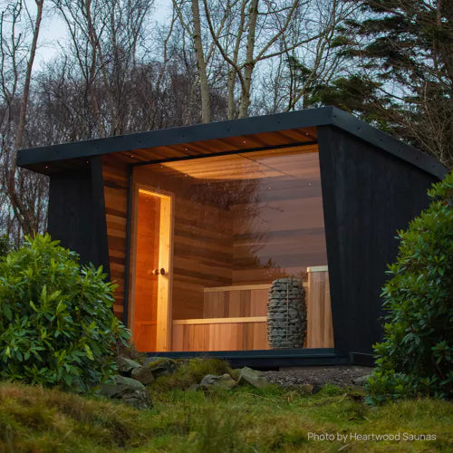 Huum Hive Mini sauna heater model HIVE Mini , located in a wooden cabin beneath a glass wall, surrounded by a serene forest environment.